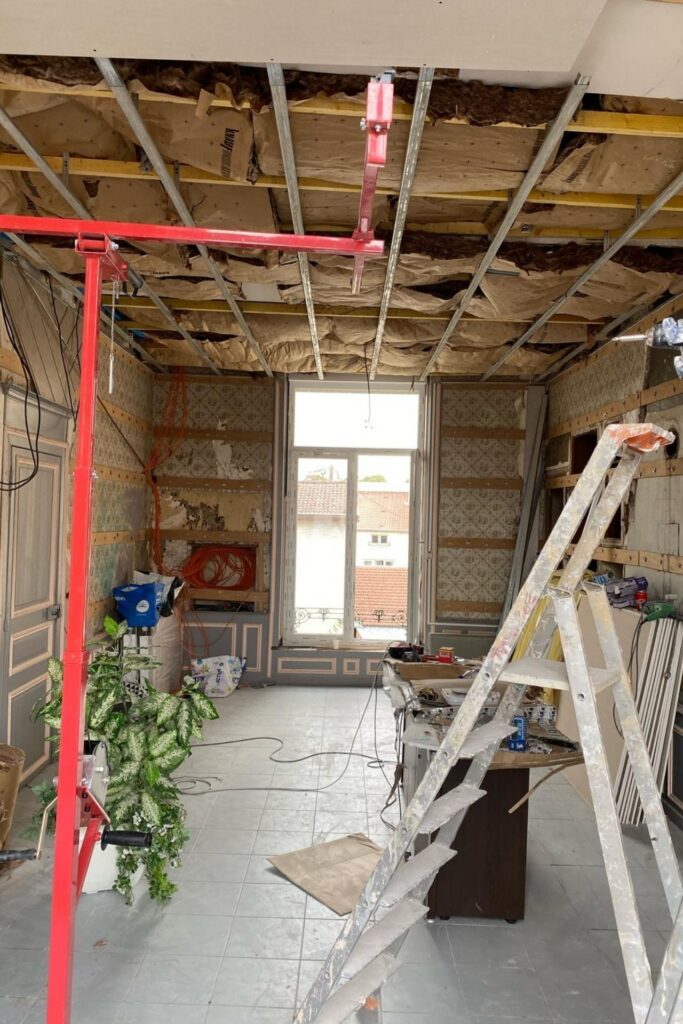 Chantier de rénovation intérieure avec plafond démoli, poutre métallique rouge, échafaudage et fenêtre double battant