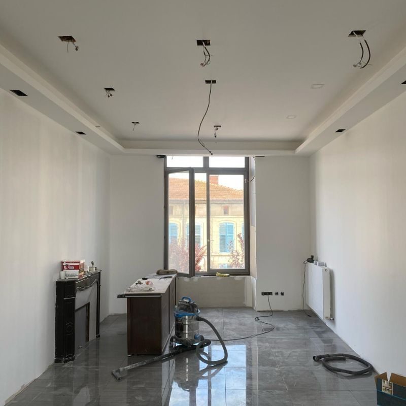 Intérieur en rénovation avec plafond blanc, installations électriques apparentes, sol gris brillant, fenêtre panoramique et